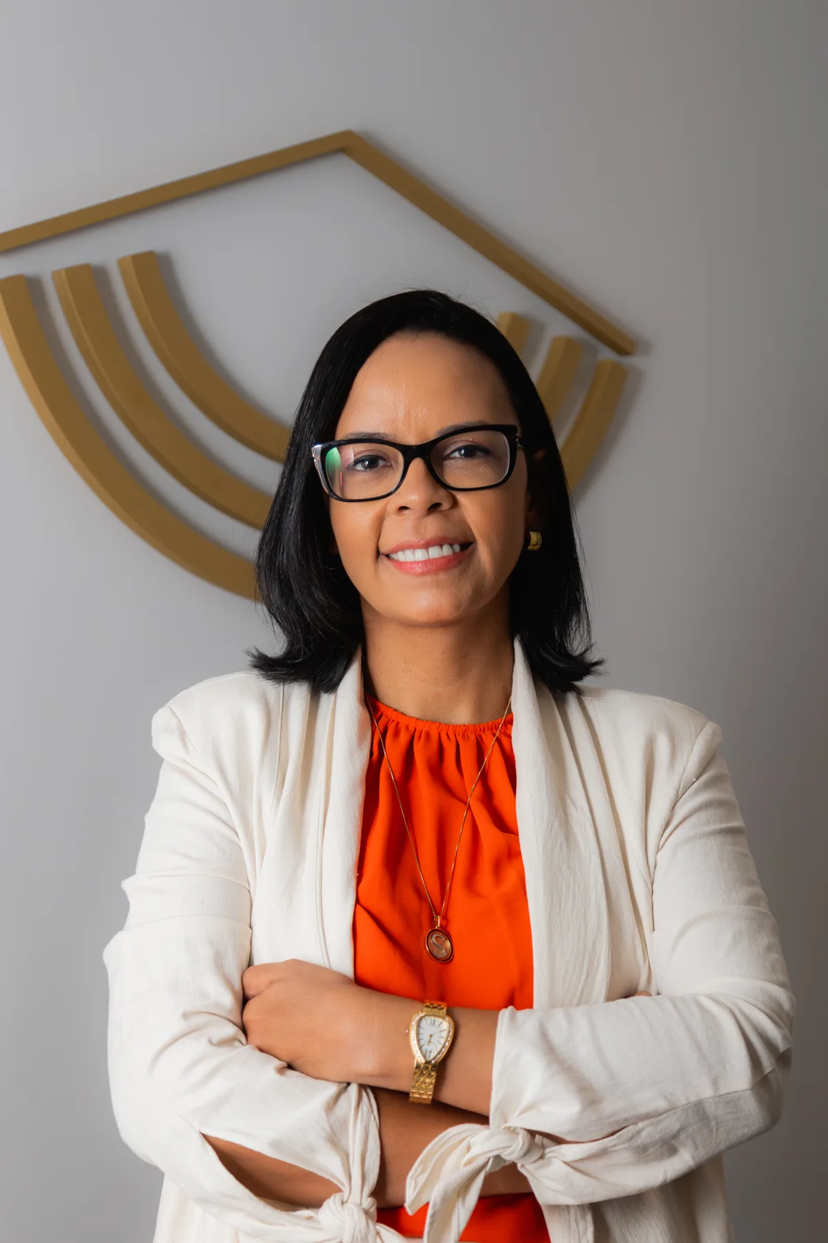 Sabrina Bueno de braços cruzados, em frente ao símbolo dourado do escritório
